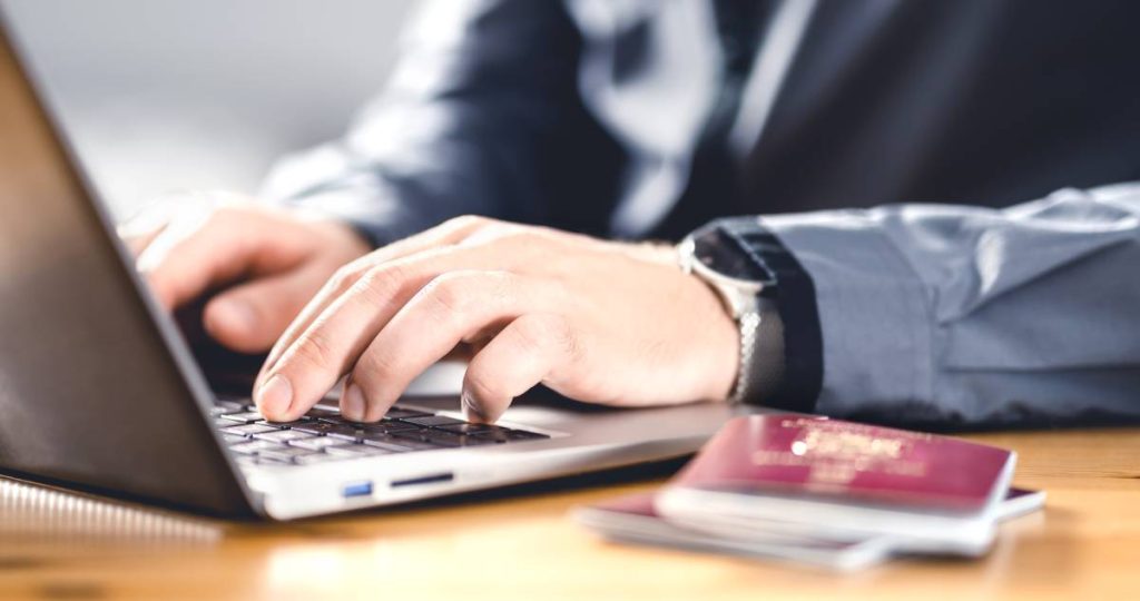 Primer plano de manos de un hombre escribiendo en un ordenador portátil con un pasaporte sobre la mesa