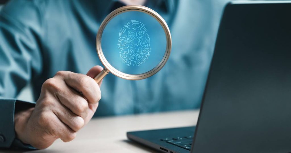 Una persona sostiene una lupa que revela una huella dactilar digitalizada frente a un ordenador portátil