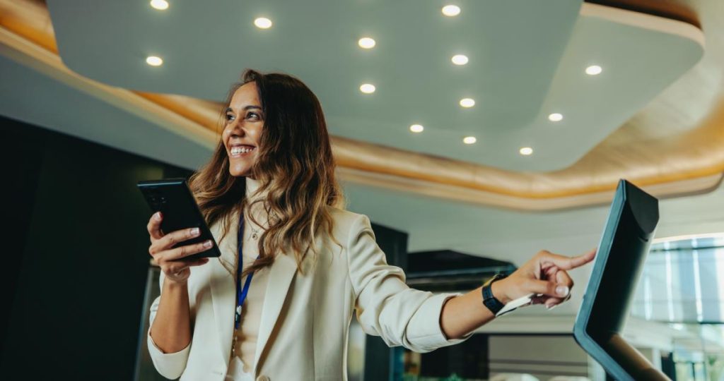Una mujer sonriente sostiene su móvil mientras interactúa con una pantalla digital táctil
