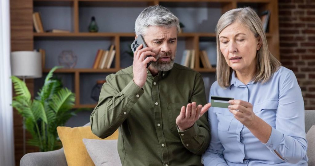 Una pareja mayor con gesto de preocupación mira una tarjeta de crédito mientras el hombre habla por teléfono
