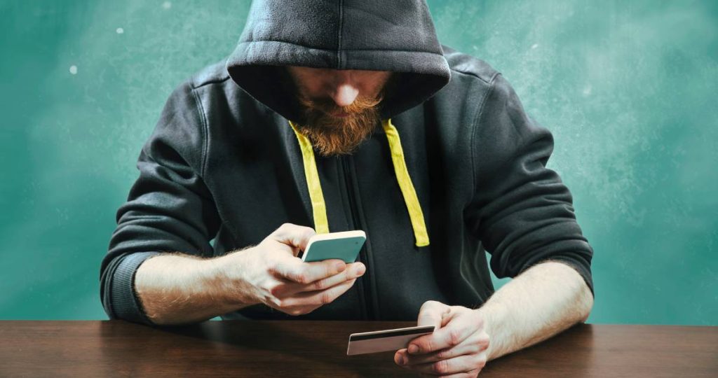 Un hombre con capucha utiliza un teléfono móvil mientras sostiene una tarjeta de crédito sobre una mesa
