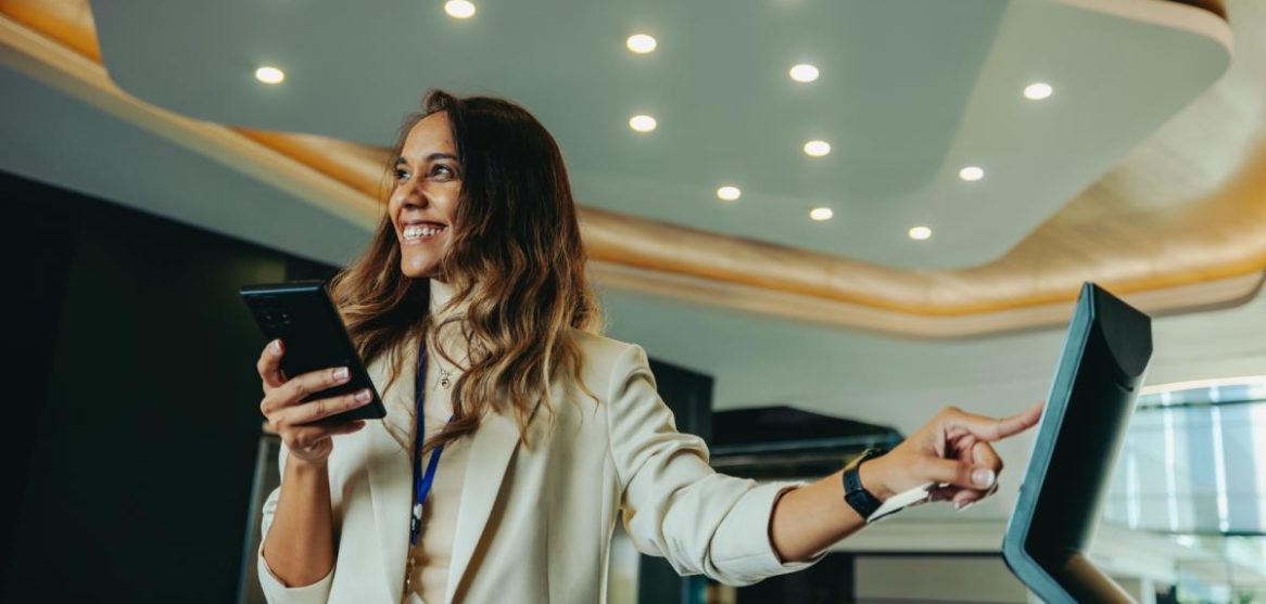 Una mujer sonriente sostiene su móvil mientras interactúa con una pantalla digital táctil