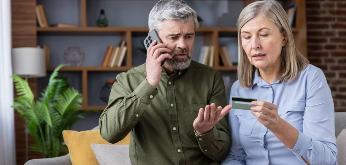 Una pareja mayor con gesto de preocupación mira una tarjeta de crédito mientras el hombre habla por teléfono
